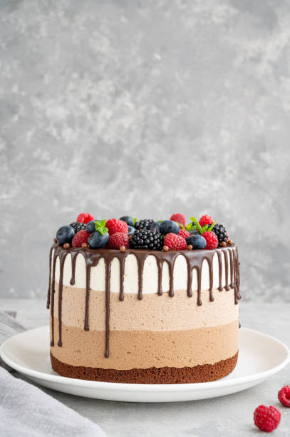 istock Three chocolate mousse cake with chocolate glaze, blueberries, raspberries and blackberries on top on a white plate on a gray concrete background. Cake with three layers of different taste. Copy space. 1341996778