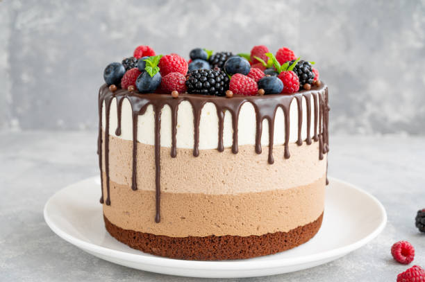 istock Three chocolate mousse cake with chocolate glaze, blueberries, raspberries and blackberries on top on a white plate on a gray concrete background. Cake with three layers of different taste. Copy space. 1341996782