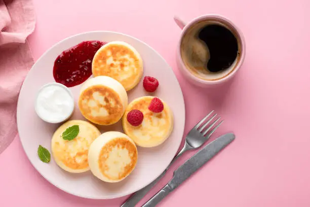 Syrniki, cottage cheese fritters with raspberry jam, yogurt and cup of coffee on pink background. Top view copy space