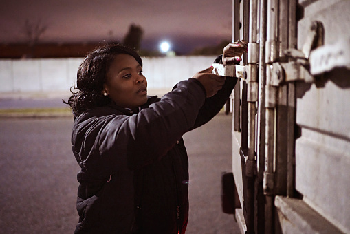 Waist-up side view of Black woman in warm clothing closing and locking rear cargo door before leaving on nighttime road trip.