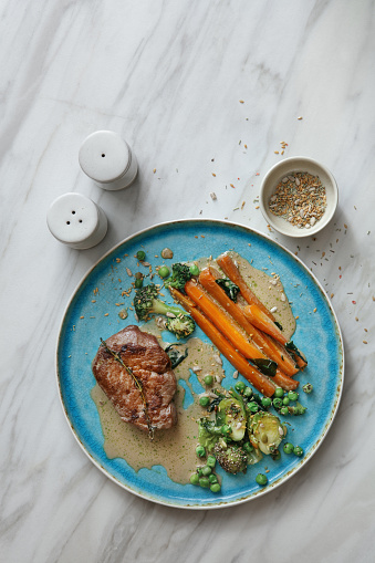 Florentine steak with glazed carrot, green pea and Brussels sprouts. Flat lay top-down composition on marble background.