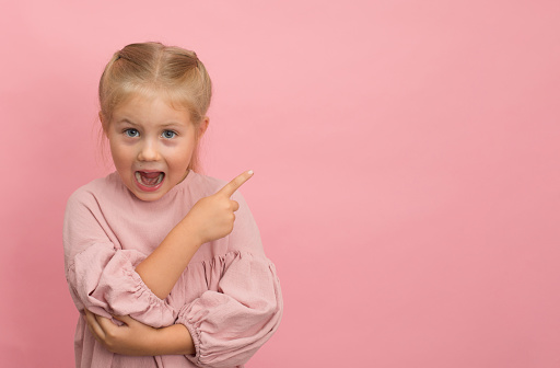 Little surprised funny girl with mouth open pointing finger aside on pink background with copy-space.