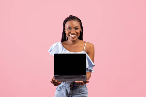 istock Funky black lady holding laptop pc with blank screen for your ad or website on pink studio background, mockup 1342027439