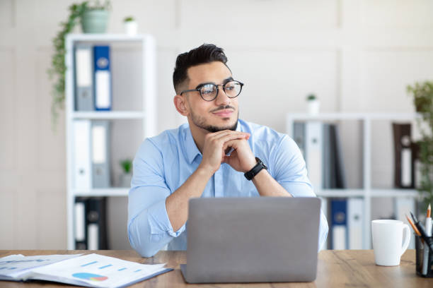 istock Dreamy Arab guy in glasses sitting at laptop at office, looking aside, taking break from online job 1342027507