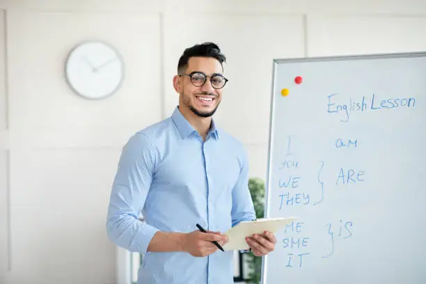 Joyful male English teacher explaining foreign language rules near blackboard, standing with clipboard, smiling at camera in office. Young Arab tutor teaching online course, giving lecture or webinar