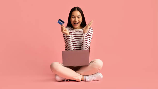 Online Payments. Cheerful young asian woman sitting with laptop and credit card Online Payments. Cheerful young asian woman sitting with laptop and credit card on floor over pink studio background, emotional excited korean female enjoying e-commerce, panorama with copy space credit card women internet currency stock pictures, royalty-free photos & images