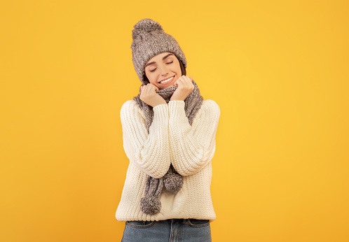Happy pretty lady wearing white knitted sweater, grey scarf and hat, standing with closed eyes over yellow studio background. Warm winter season fashion accessories concept