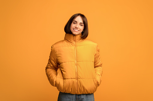 Ropa de invierno. Joven mujer sonriente con chaqueta abrigada, tomados de la mano en los bolsillos y mirando a la cámara, fondo amarillo photo