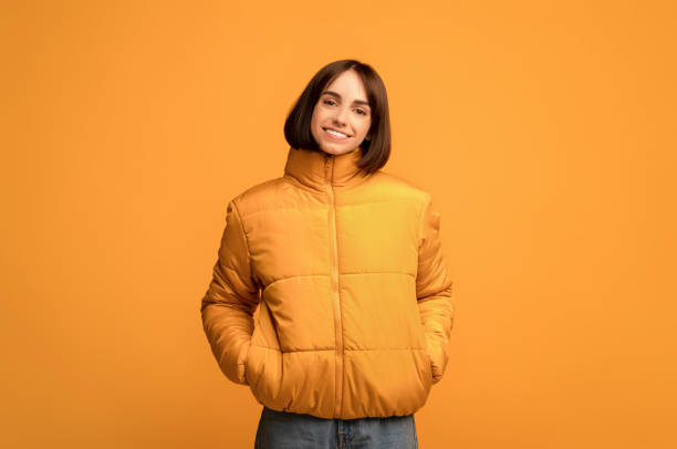 Winter clothing. Young smiling woman in warm jacket, holding hands in pockets and looking at camera, yellow background Winter clothing. Young smiling woman in warm jacket, holding hands in pockets and looking at camera standing over yellow studio background. Warm clothes fashion concept jacket stock pictures, royalty-free photos & images