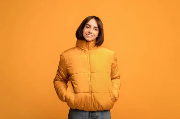 Photo of Winter clothing. Young smiling woman in warm jacket, holding hands in pockets and looking at camera, yellow background