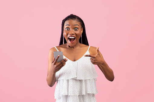 Shocked black lady pointing at smartphone in excitement, winning lottery or online casino bet on pink studio background. Excited African American woman receiving amazing news on phone