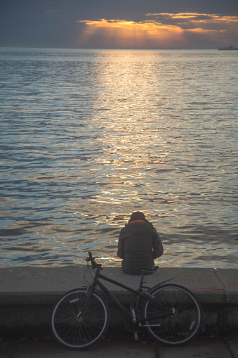 Istanbul, Turkey - December 9, 2020: Lonely teenager and his bike by the sea at sunset. Back view, vertical shot. Break, city life and loneliness concepts.