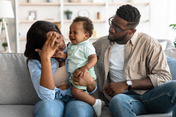 Tired African American parents sitting with crying kid on sofa Portrait of tired black mum sitting with her small crying black kid on couch at home, feeling headache and migraine, touching forehead with hand, sad dad comforting her. Exhaustion And Parenthood postpartum depression stock pictures, royalty-free photos & images