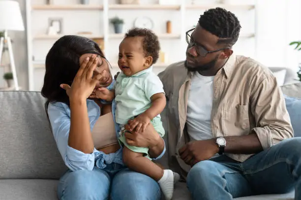 Photo of Tired African American parents sitting with crying kid on sofa