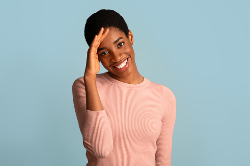 Portrait Of Shy Millennial African American Woman Smiling And Touching Head In Embarrassing, Cute Young Black Female Holding Hand Near Forehead, Posing Over Blue Studio Background, Copy Space