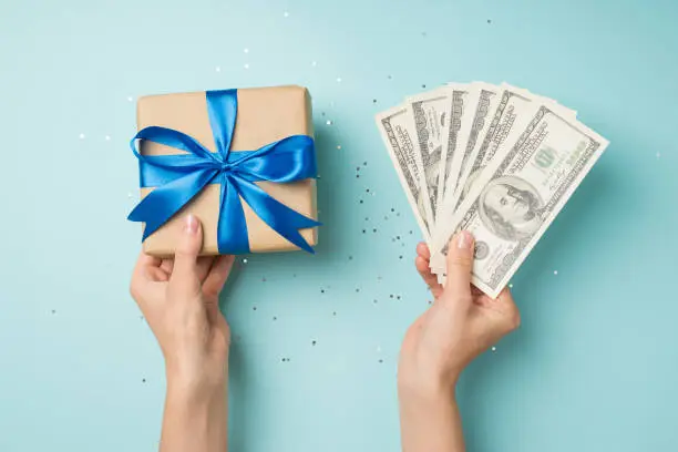 Photo of First person top view photo of hands holding fan of money and craft paper giftbox with blue ribbon bow over shiny sequins on isolated pastel blue background