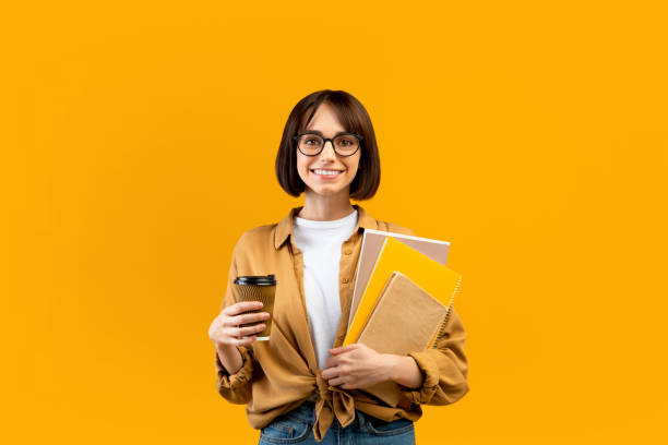 istock Happy female student holding notebooks and coffee to go, having break while studying, posing over yellow background 1342037670