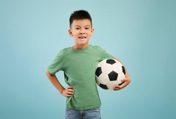 bambino asiatico felice con pallone da calcio in mano su sfondo blu - soccer child indoors little boys foto e immagini stock