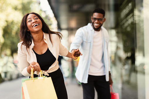 Sales Season. Portrait Of Smiling Excited African American Lady Pulling Boyfriend To Shopping Store To Buy New Clothes, Fooling And Running To Mall Together. Discount, Fun, Purchase And Retail