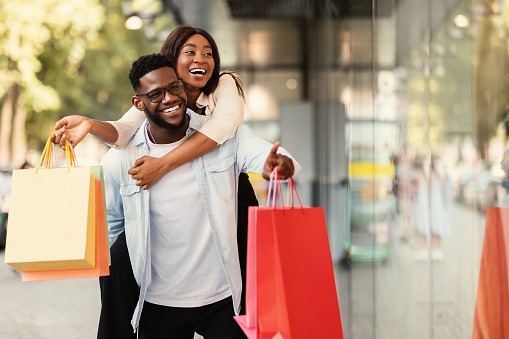 Portrait of smiling African American man giving piggyback ride to his happy woman who holding shopper bags, couple looking and pointing at shop window, walking near shopping center