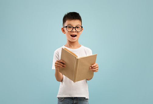 Excited little asian boy wearing eyeglasses holding and reading open book, cute schoolboy in spectacles enjoying study and learning, standing isolated over blue studio background, copy space