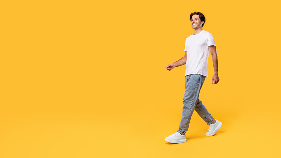 Advertising And Marketing. Full length body size profile side view of nice cheerful bearded guy in casual white t-shirt and jeans walking looking at free empty space isolated over orange color wall