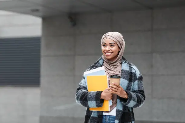 Photo of Happy pretty cute afro american female student in hijab and coat with books and cup of coffee ready to study