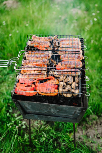 asa salchichas con verduras y especias en un brasero. vista superior. surtido de deliciosa carne a la parrilla en parrilla barbacoa con humo y llamas en hierba verde. - vertical meadow mushroom vegetable fotografías e imágenes de stock