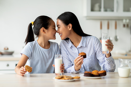 Loving asian mother and little daughter having snacks and drinking milk in kitchen interior, enjoying freshly baked cakes. Happy family eating homemade pastry and bonding at home