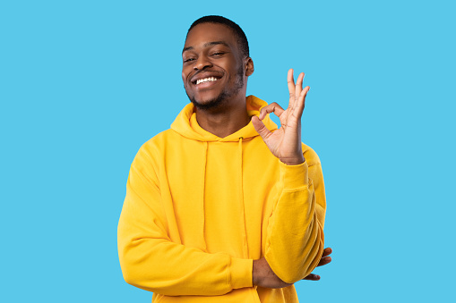 I'm Okay. Cheerful Black Man In Yellow Hoodie Gesturing OK Sign Smiling To Camera Standing Over Blue Studio Background. Portrait Of Contented Customer Approving Something