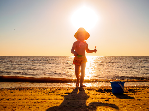 Silhouette of a little girl in a panama hat on the seashore against the backdrop of the setting sun. A girl plays with a toy spatula and a bucket in the sand.