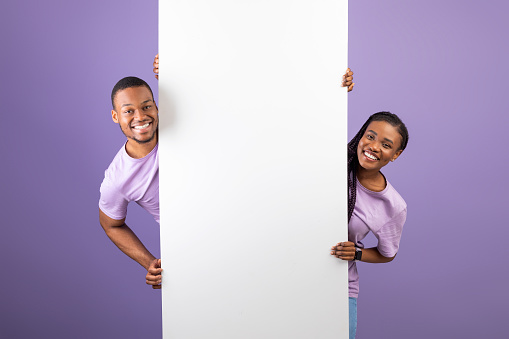 Smiling young black couple holding white vertical advertisement board, demonstrating free copy space for your text or design, positive guy and lady peeking out banner, purple violet background