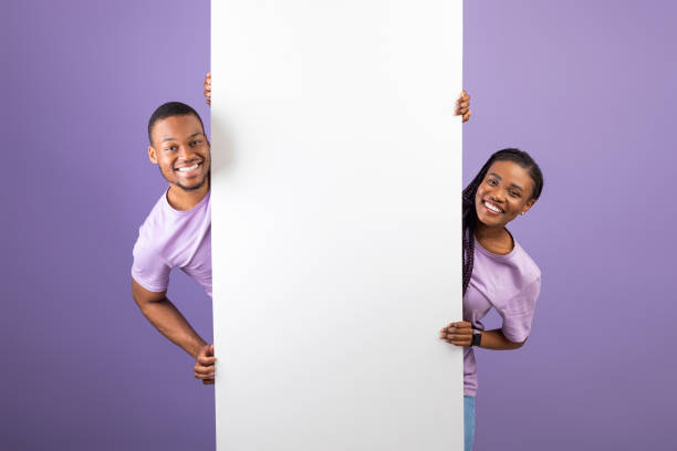 Black couple holding and pointing at blank white advertising placard Smiling young black couple holding white vertical advertisement board, demonstrating free copy space for your text or design, positive guy and lady peeking out banner, purple violet background hiding place stock pictures, royalty-free photos & images