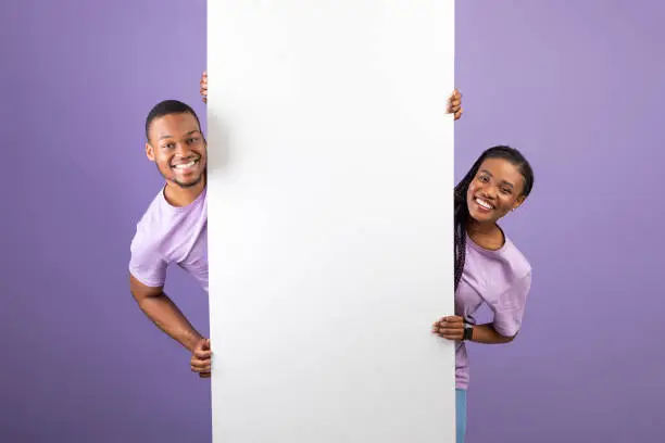 Smiling young black couple holding white vertical advertisement board, demonstrating free copy space for your text or design, positive guy and lady peeking out banner, purple violet background