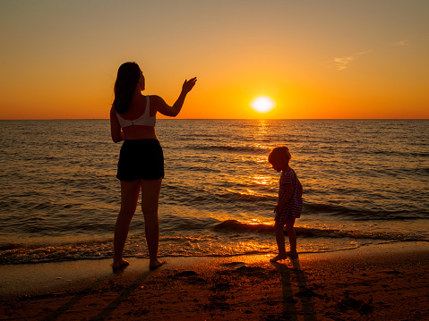 Mom and little daughter are standing side by side on the seashore against the backdrop of the setting sun.