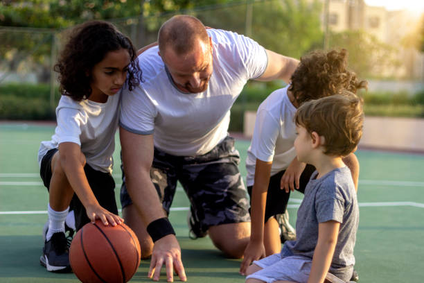 Mixed age group of school children listen their coach as he explains the next play. Shot of three young boys and their coach chatting outside on basketball court. Mixed age group of school children listen their coach as he explains the next play. Young coach giving his small team talk. youth sports competition stock pictures, royalty-free photos & images