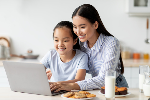Happy asian mother and kid using laptop together, sitting at kitchen table and looking at computer screen, eating homemade pastry. Family spending time together at home