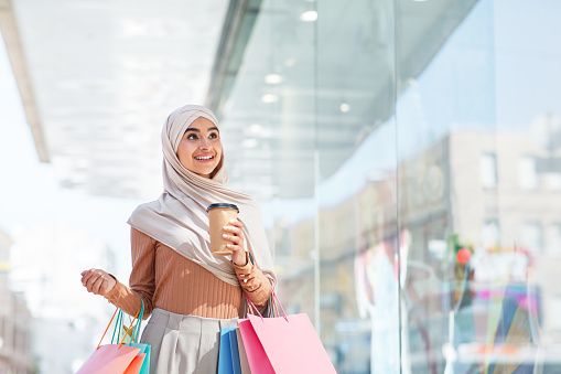 Inspired happy emotional cute millennial middle eastern lady in hijab with many packages and cup of drink looks in shop windows near mall. Great discount, huge sale, joy with shopping, positive mood