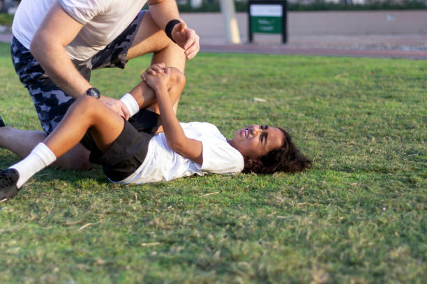 Shot of man looking at injured boy and trying to help him. Young sports boy with injured knee getting help from man touching his knee. Injured boy lying on the grass in pain, making face expression. Shot of man looking at injured boy and trying to help him. knee to the head pose stock pictures, royalty-free photos & images