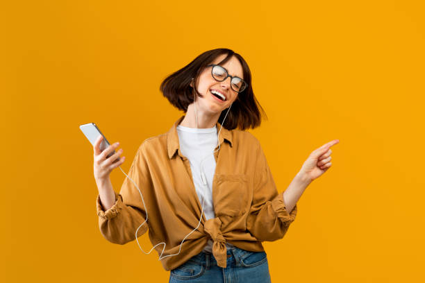 ótima canção. mulher animada ouvindo música em fones de ouvido no smartphone, dançando e se divertindo em fundo amarelo - music headphones women singing - fotografias e filmes do acervo