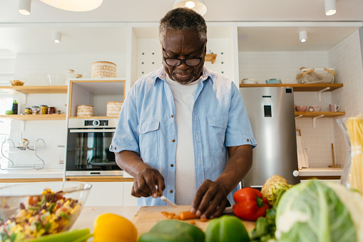 Senior African man standing in the kitchen and chopping carrot on wooden board. He is preparing a healthy dinner.