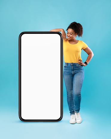 Joyful Afro woman in casual wear leaning on huge cellphone with empty white screen on blue studio background, mockup for mobile app, website, your ad design. Smartphone template