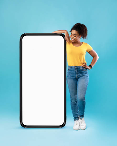 mulher afro alegre em casual desgaste inclinando-se em celular enorme com tela branca vazia no fundo azul, mockup - ecrã grande - fotografias e filmes do acervo