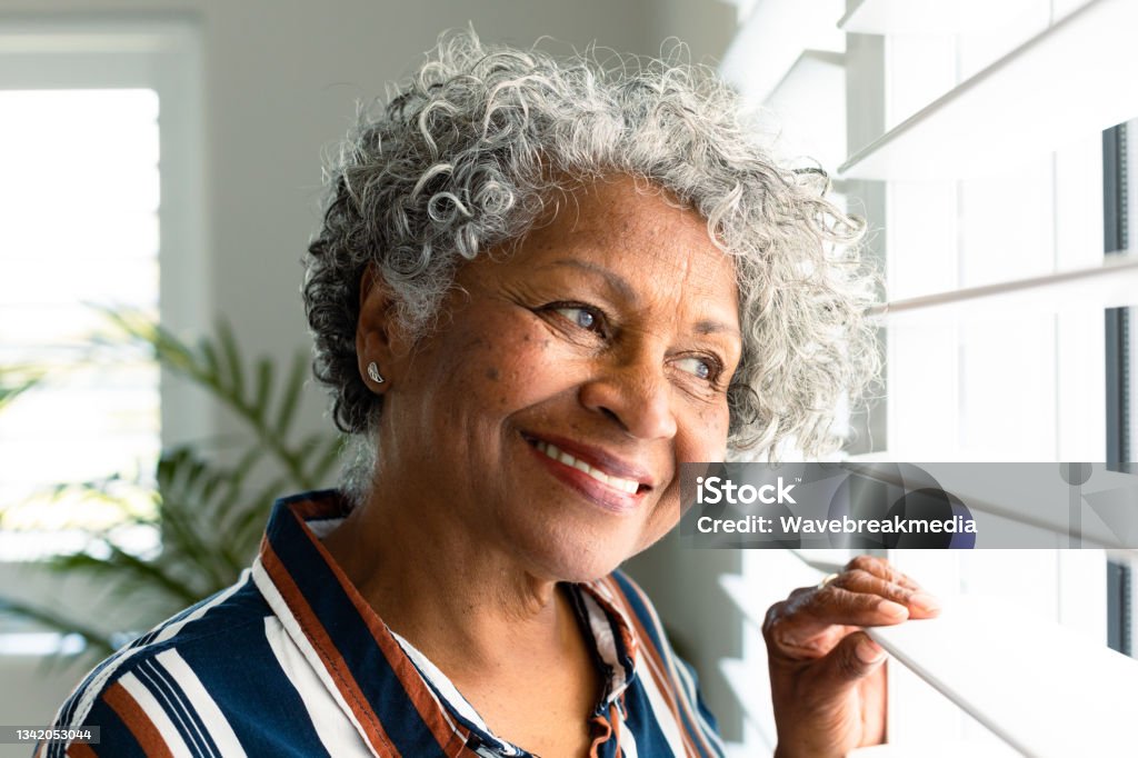 Portrait of smiling african american senior woman looking through window Portrait of smiling african american senior woman looking through window. healthy retirement lifestyle at home. Senior Women Stock Photo