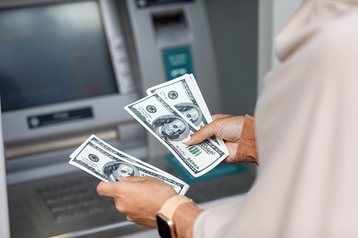 Hands of unrecognizable young arabian business woman in hijab counts dollars near atm with empty screen, cropped. Online payment, money transfers, financial transactions and digital financial services