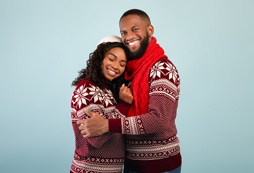 Loving happy african american spouses in Christmas sweaters hugging and smiling to camera, standing over blue studio background. Expressing affection.