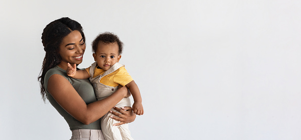 Motherhood Concept. Portrait Of Smiling Young Black Mother Holding Adorable Infant Son On Arms, Happy African Mom Enjoying Time With Her Baby, Standing Over White Background, Panorama With Copy Space