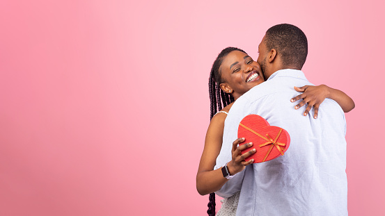 Passionate black lovers hugging on pink background, lady holding heart shaped gift box. Affectionate couple celebrating Valentine's Day or anniversary together, banner, panorama, free copy space