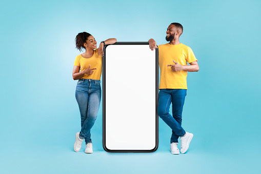 Hot offer, online sale. Funky black young couple pointing at cellphone with mockup space over blue studio background. Afro guy and lady advertising new awesome mobile app or cool website