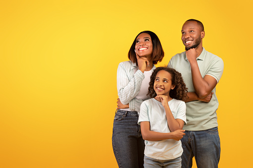Special offer. Young black family of three looking aside at free space and holding hands on chin, standing over yellow background, studio shot, banner
